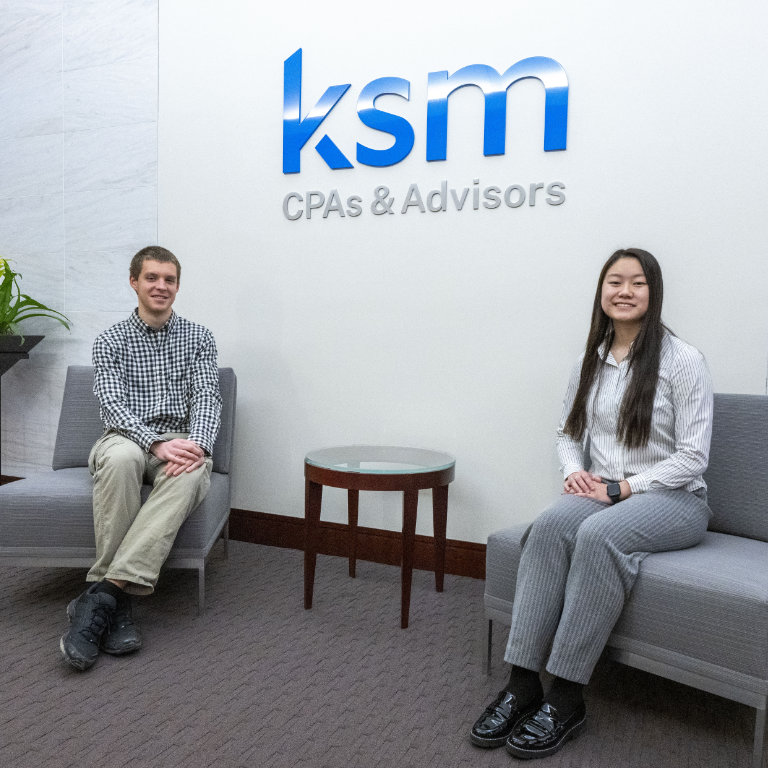 Two student interns sit on chairs under the KSM CPAs & Advisors office logo. 