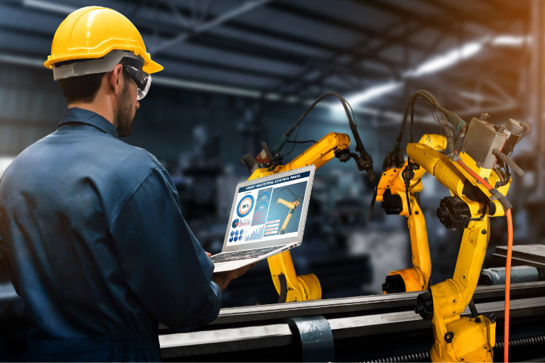 A worker in a hard hat looks at a screen next to robotic arms on a conveyer belt. 