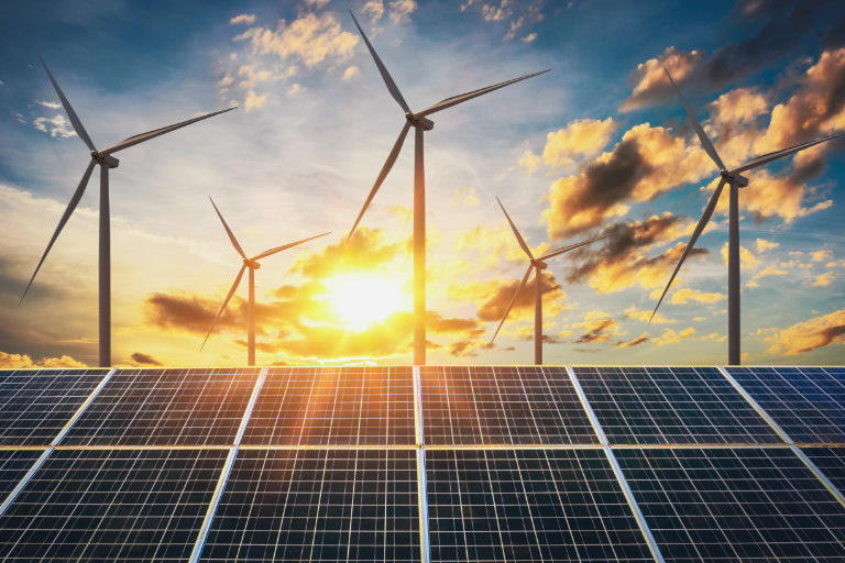 Wind turbines turn in front of a solar panel at sunset.