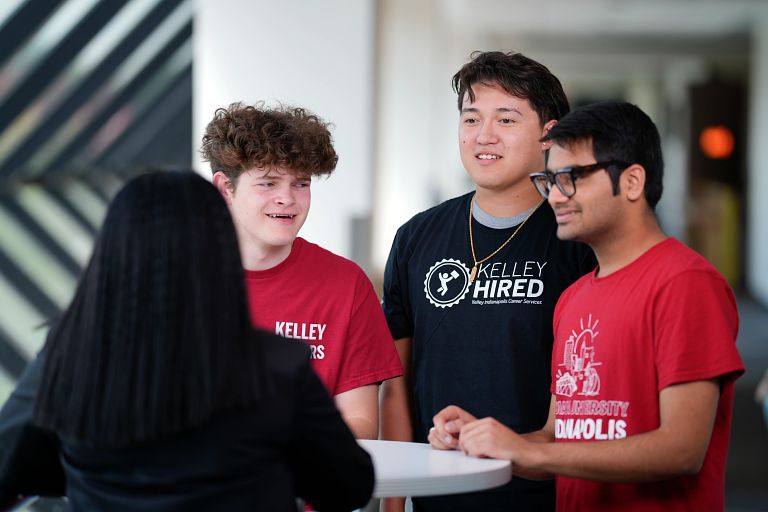 Three students chat with a professor at a table in a Kelley hallway. 